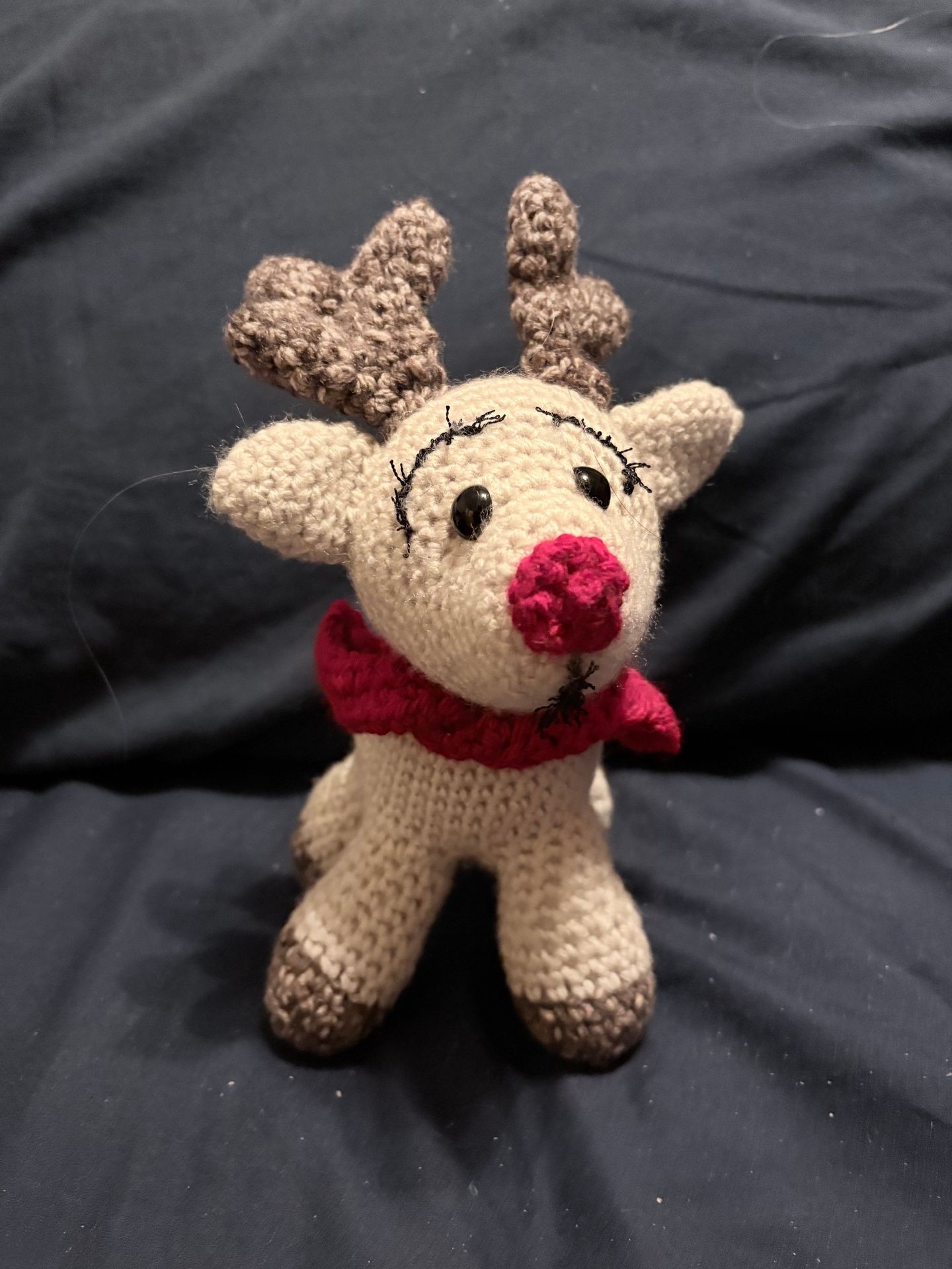 Knit reindeer toy with red nose and scarf on dark fabric background.
