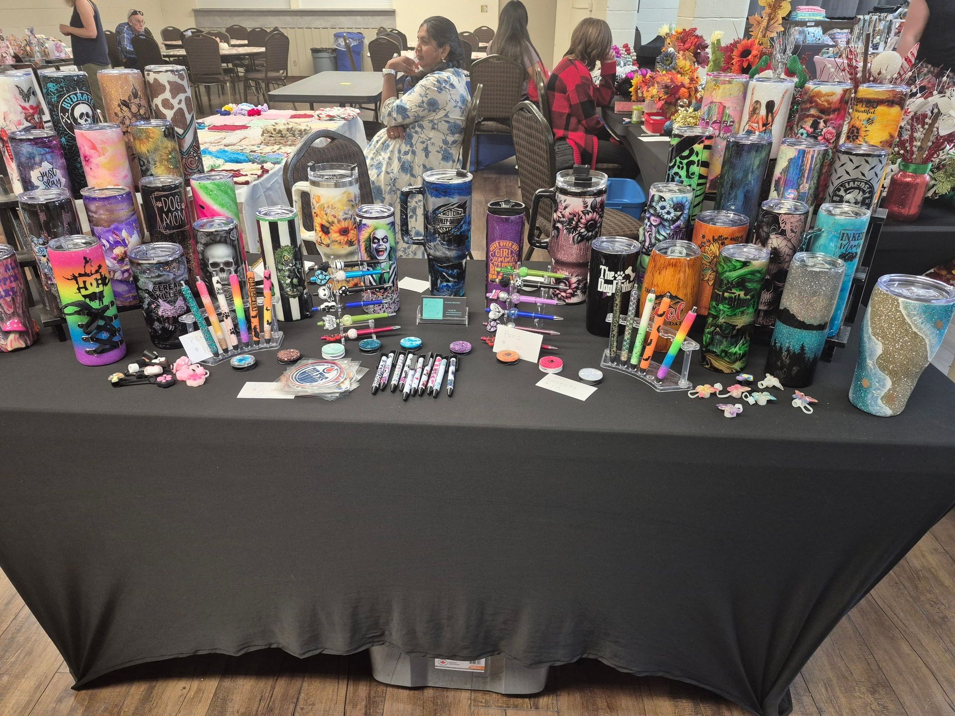 Table displaying various custom tumblers and pens with colorful designs in a craft fair setting.